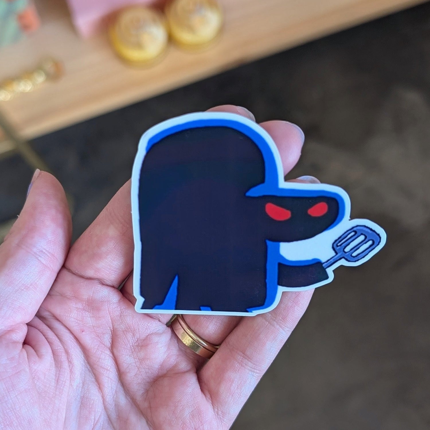 Hand holding a dark blue ghost-shaped sticker with red eyes on a wooden shelf background.