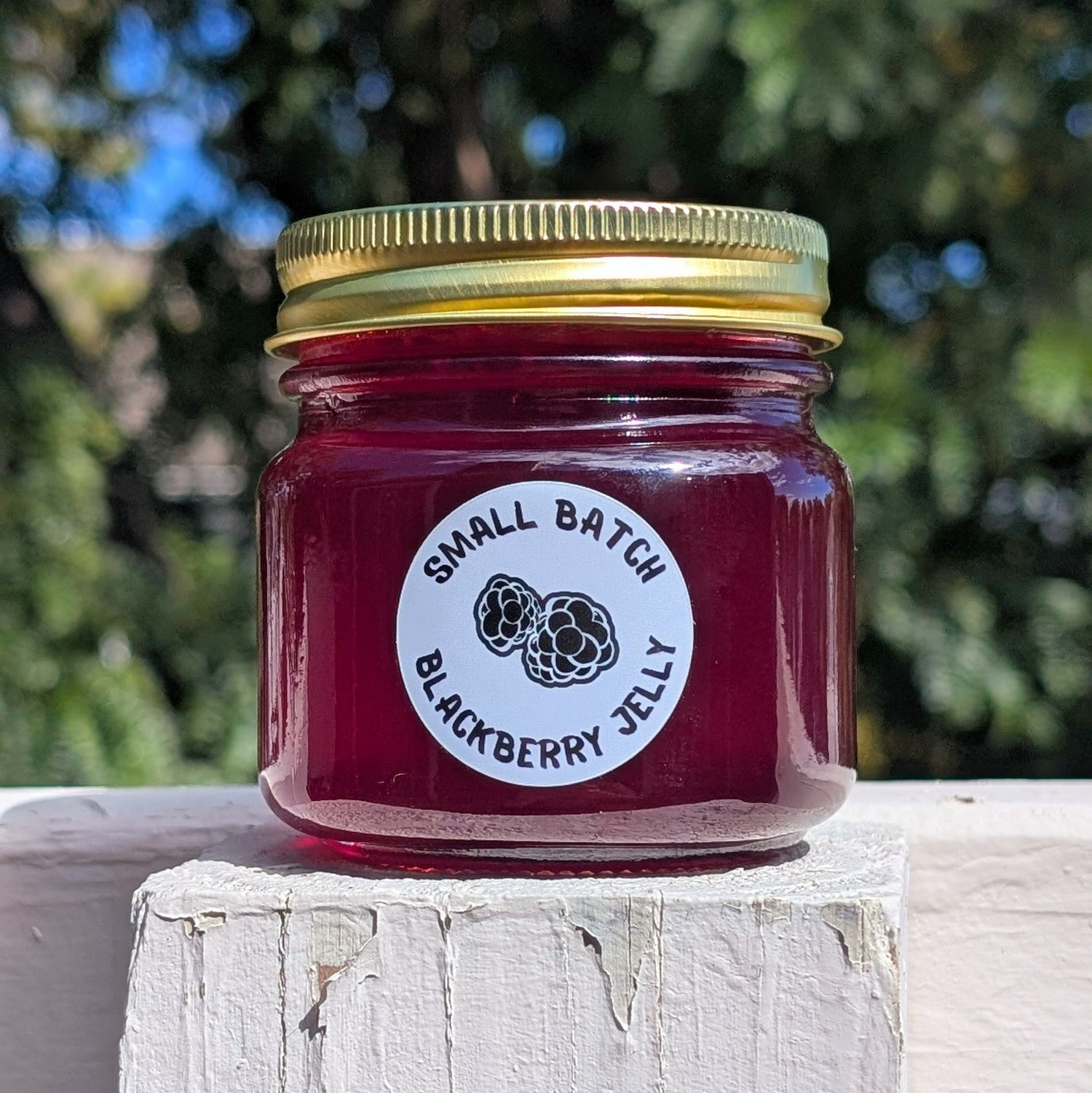Jar of small batch blackberry jelly with a gold lid on a white wooden surface outdoors in front of a blurred out green tree background.