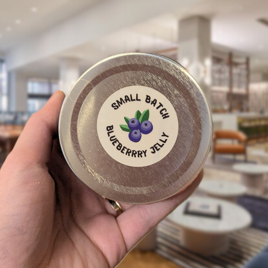 Small batch blueberry jelly jar lid held by a hand with blurred hotel lobby in the background.