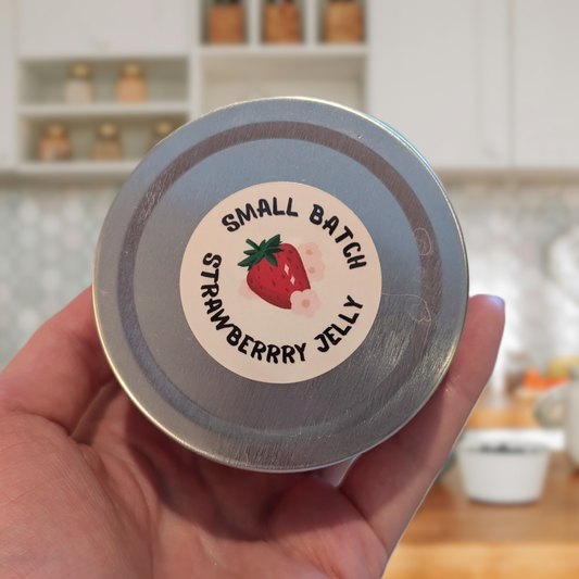 Hand holding a jar lid labeled 'Small Batch Strawberry Jelly' with a blurred kitchen background.
