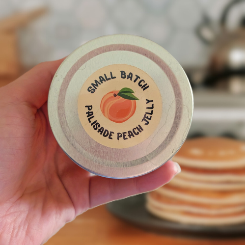 Hand holding a jar of palisade peach jelly with a kitchen background featuring pancakes.